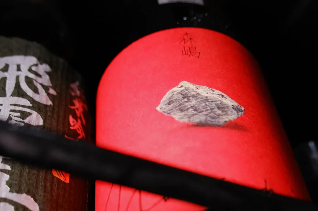 A close-up of a red label featuring a rock illustration, partially obscured by dark shadowy elements and surrounded by other bottles with decorative labels.