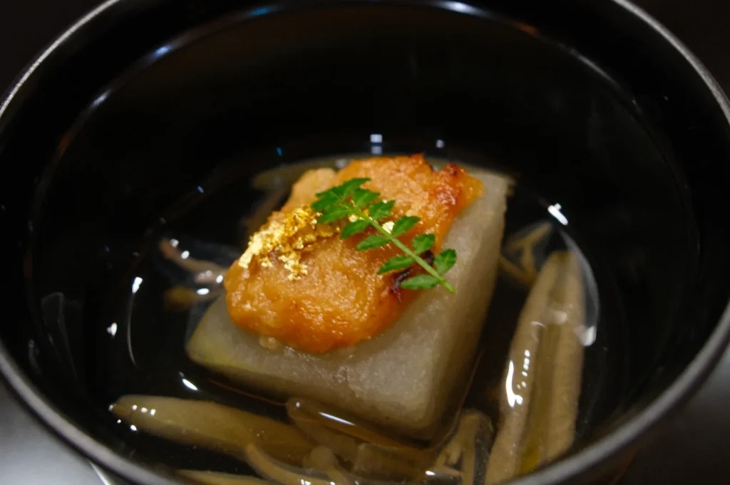 A delicate Japanese dish in a black bowl features a golden-topped daikon radish with a sprig of green leaf and gold flakes, nestled in a clear broth.