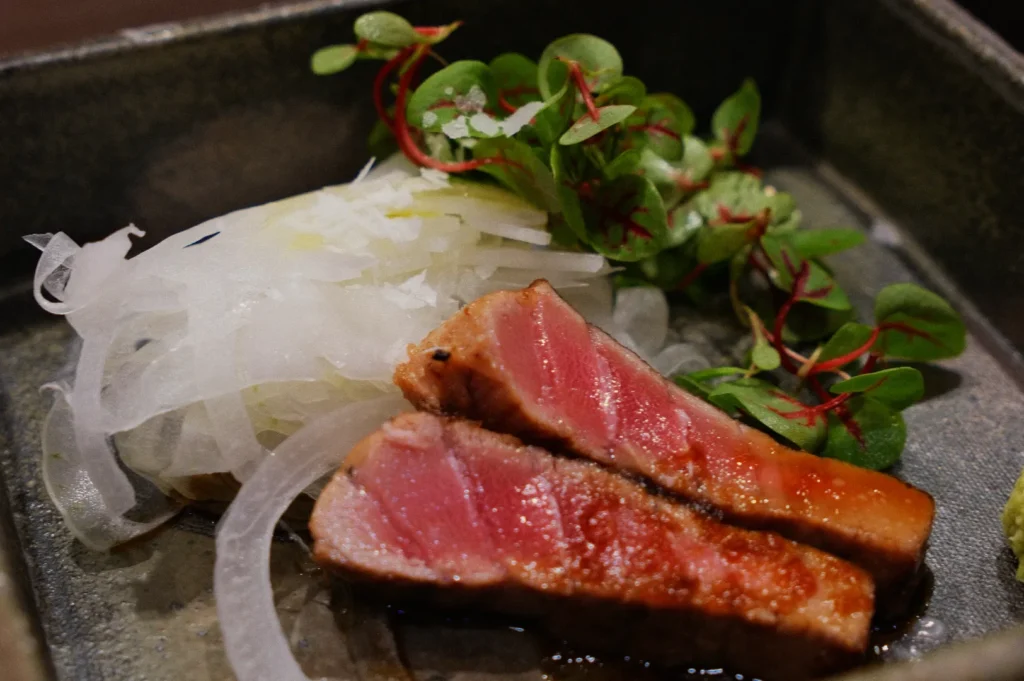 A dish featuring two slices of seared meat, accompanied by thinly sliced onions and fresh greens.