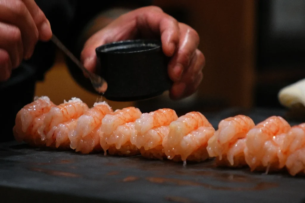 Close-up of a chef's hands delicately sprinkling seasoning from a small bowl onto a row of raw shrimp on a dark counter, evoking a sense of precision.