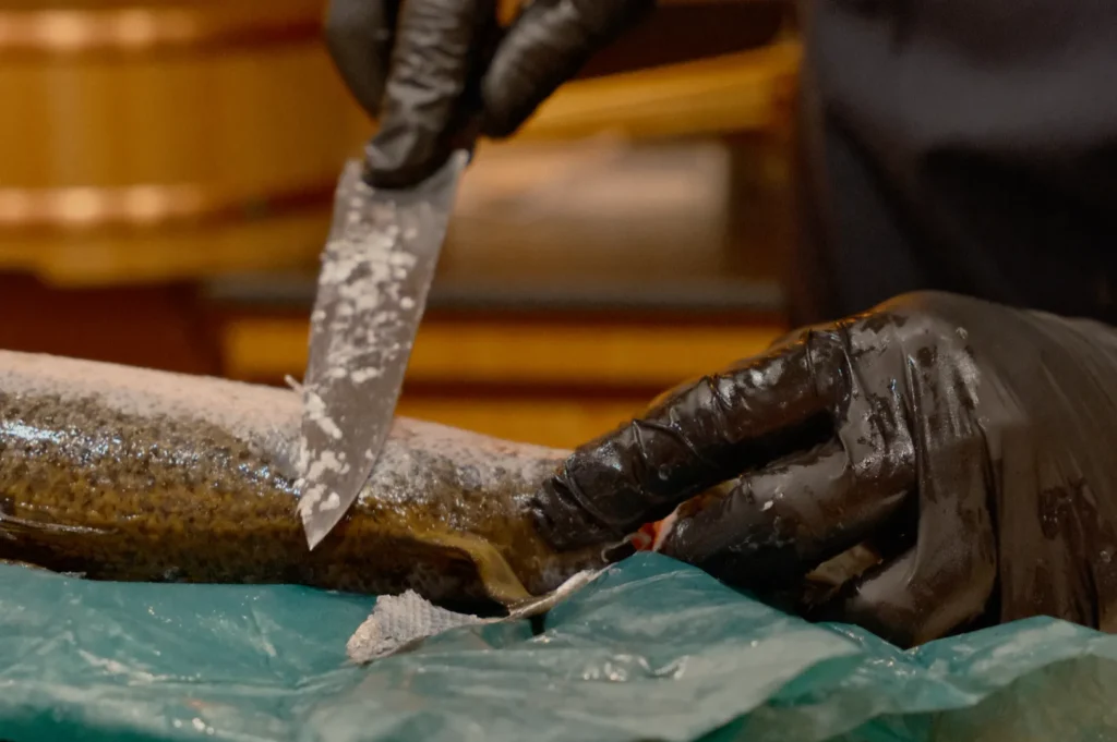 Gloved hands fillet a fish with a sharp knife on a blue cutting surface. The setting appears to be a kitchen, with an emphasis on precision and skill.