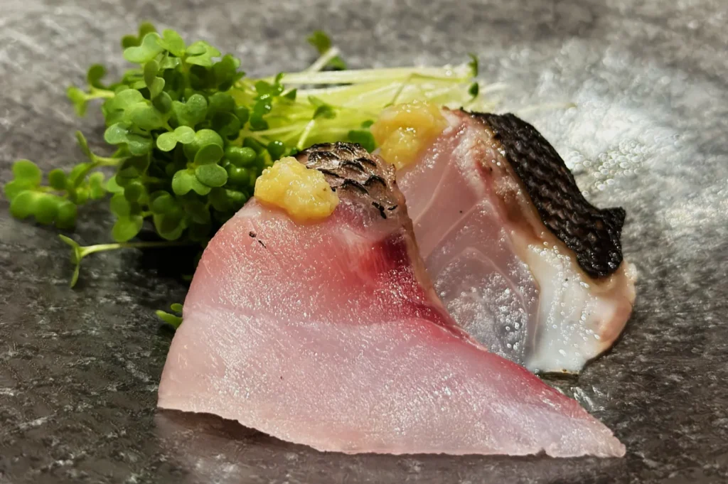 Close-up of two slices of sashimi topped with a small amount of grated ginger, garnished with fresh microgreens. The scene has a fresh, appetizing look.
