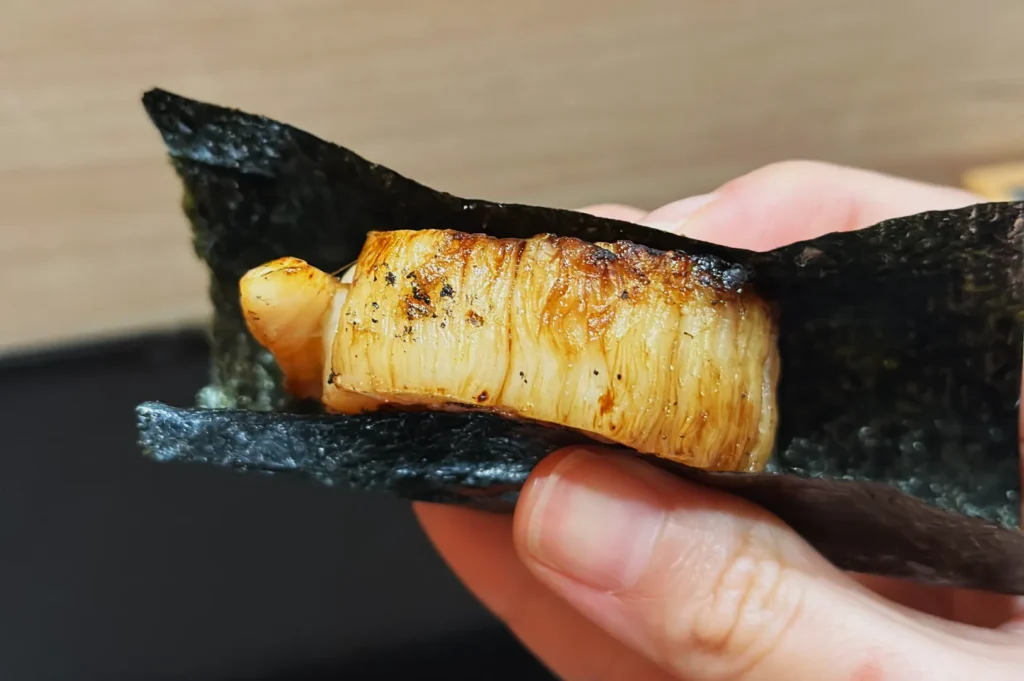 A hand holds a grilled scallop wrapped in a piece of dark green seaweed, displaying its golden-brown, slightly charred texture. The setting has a warm, inviting tone.