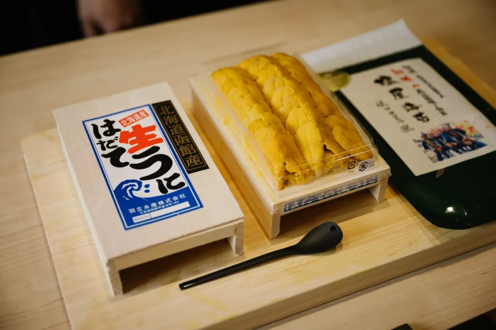 A package of fresh sea urchin with a small spoon, placed on a wooden surface next to an additional item.