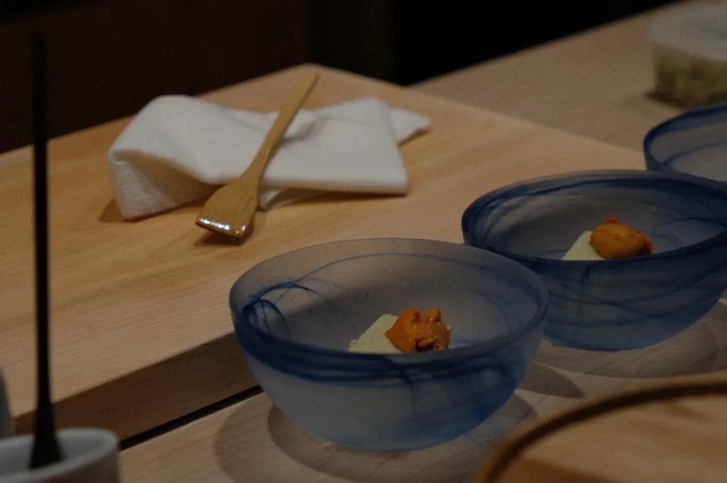 Three delicate blue bowls with sea urchin and tofu on a wooden counter, next to a folded white napkin and a bamboo utensil, create an elegant and minimalist presentation.