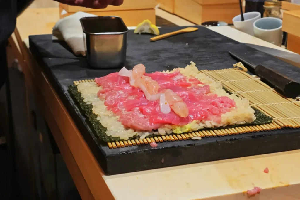 A sushi preparation surface with a sheet of nori and various types of fish laid out on top of sushi rice.