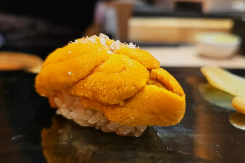 A piece of sushi topped with bright orange uni (sea urchin) on a bed of rice, garnished with a sprinkle of salt.