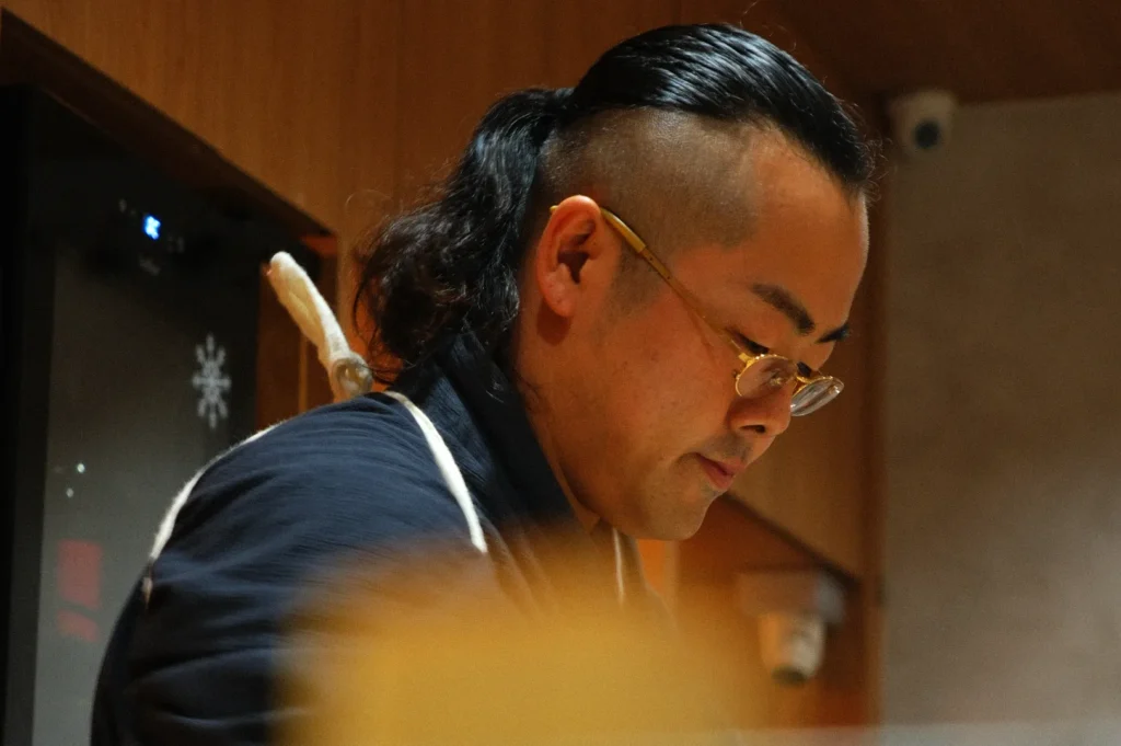 A focused chef with glasses and a shaved-side hairstyle works intently in a warm-lit kitchen, exuding concentration and precision, clad in a dark apron.