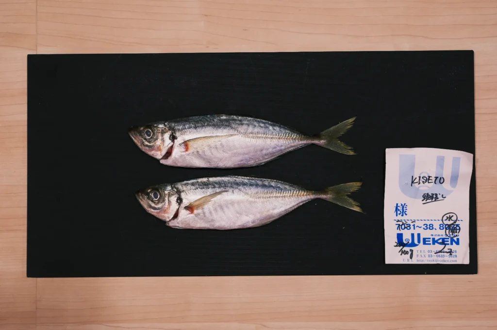 Two raw fish are laid side by side on a black cutting board, next to a label with Japanese text. The scene is minimalistic and orderly.