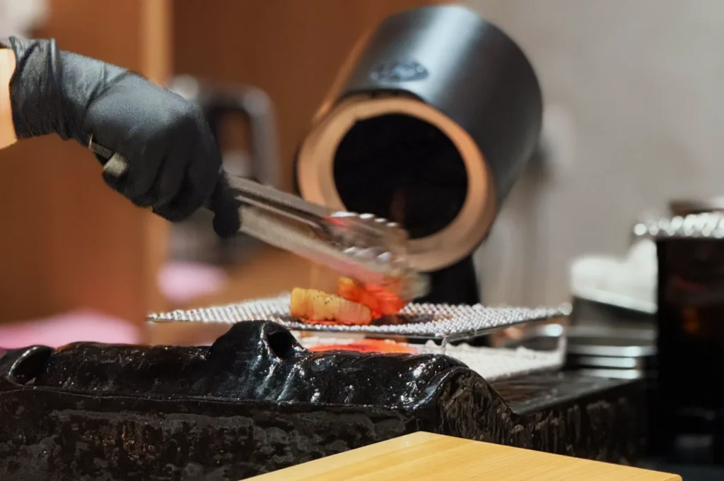 A hand wearing a black glove using tongs to place food on a grill in a cooking setup.