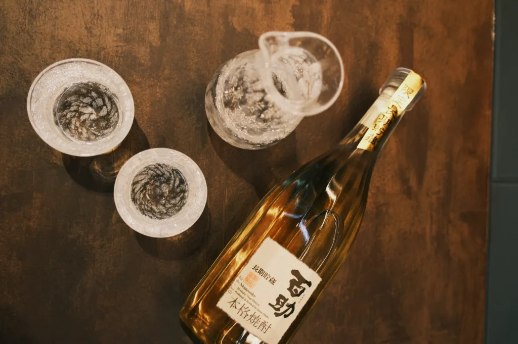 A bottle of sake on a wooden table, accompanied by two frosted sake glasses and a small pitcher.