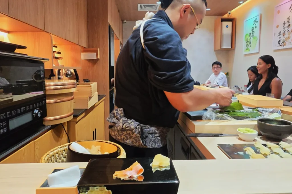 Chef Masa preparing sushi in a restaurant kitchen, with patrons seated at the counter in the background.