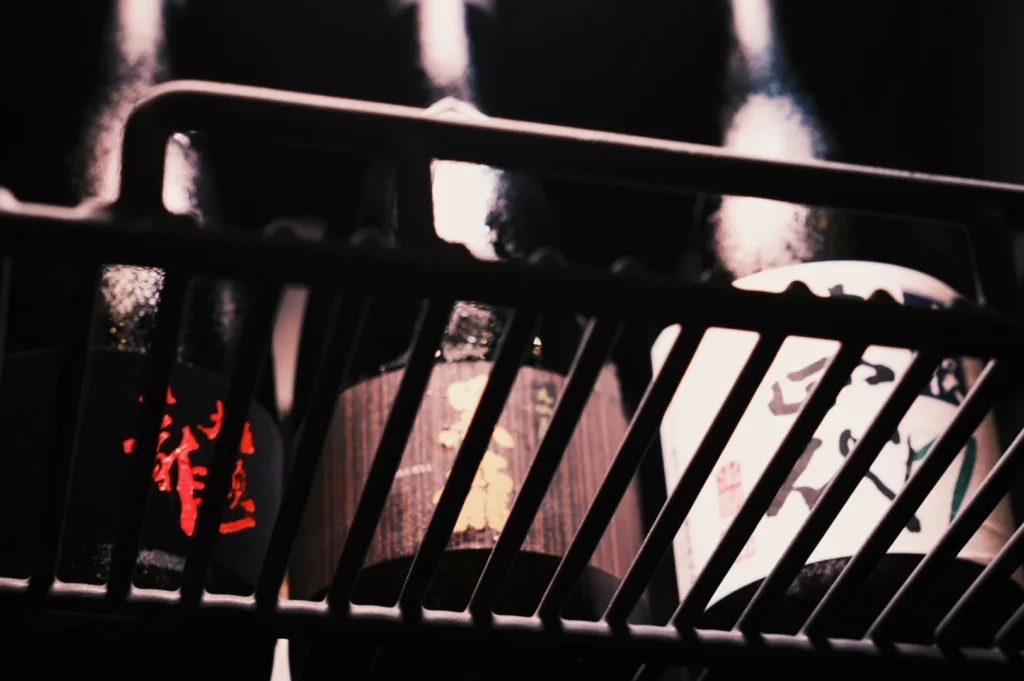 Close-up of three sake bottles viewed from below, behind a metal rack. Labels feature Japanese characters in red, gold, and black on a muted background.