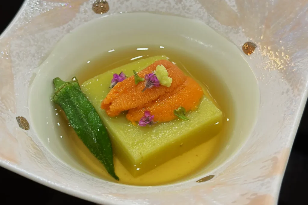 A delicate Japanese dish features a green tofu-like square topped with vibrant orange sea urchin, tiny purple flowers, and a dollop of wasabi, accompanied by an okra on the side, all in a light broth.