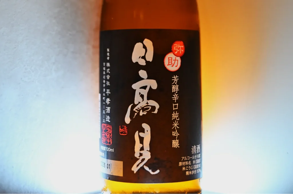 A close-up of a bottle label featuring Japanese text, indicating it is a type of sake with a design that includes a logo and various characters. The label has a dark background with white text and some red accents. The bottle appears to be illuminated from behind, creating a glow effect.