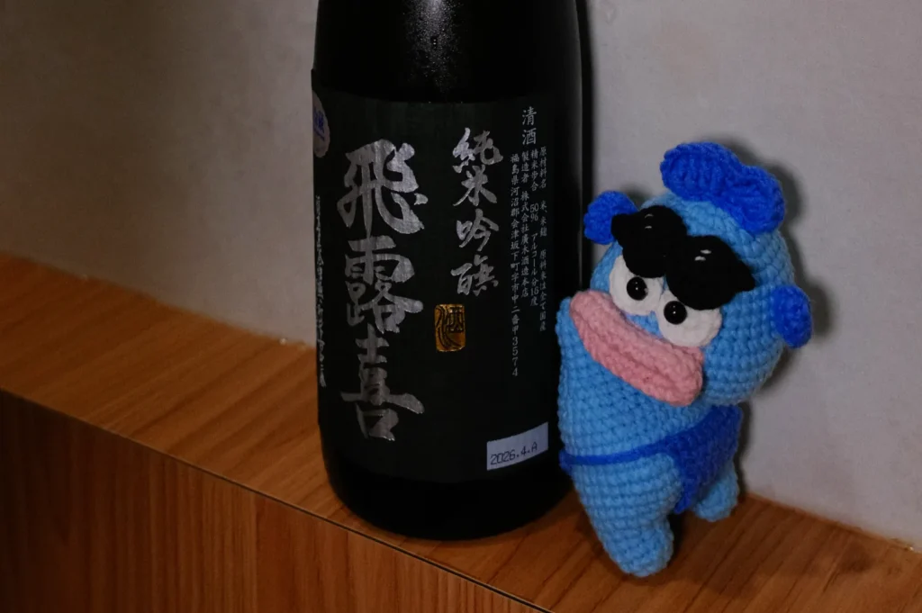 A crocheted blue character with sunglasses next to a bottle of sake with Japanese text on the label.