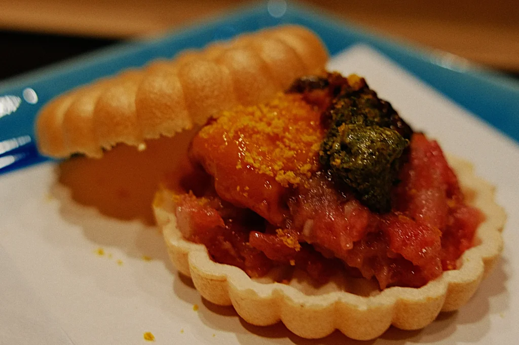 A delicate tart shell filled with vibrant, raw minced seafood topped with orange roe and green garnish, presented on a blue plate, conveying freshness and elegance.
