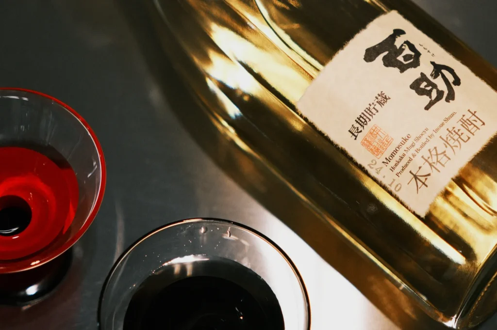A bottle of sake next to two small glasses, one containing red liquid and the other containing black liquid.