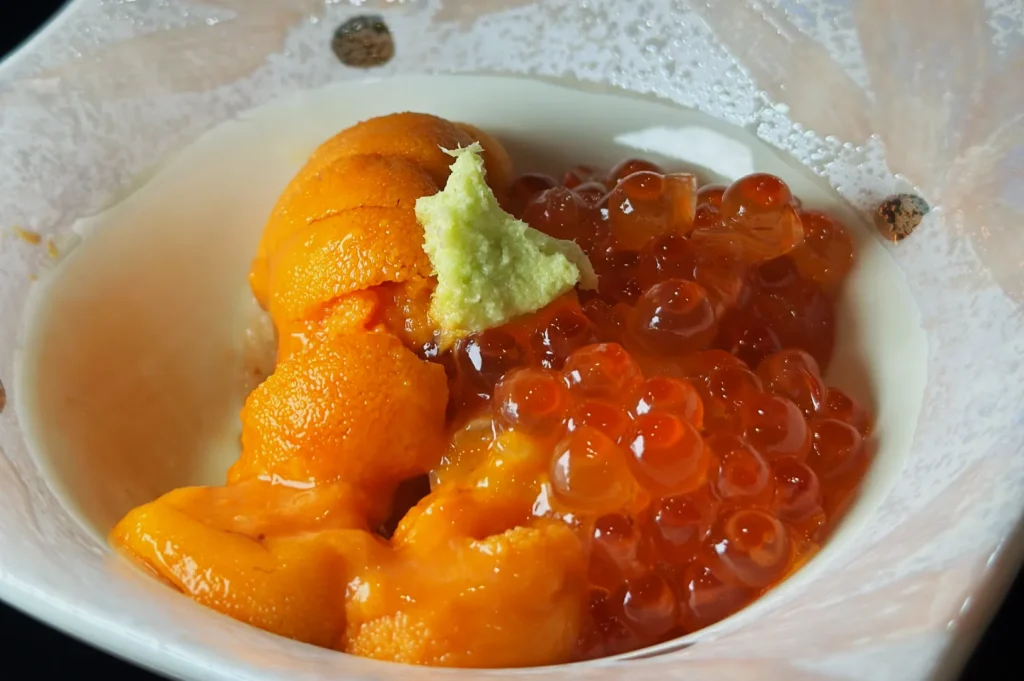A bowl containing orange sea urchin (uni) topped with bright orange fish roe (ikura) and a small dollop of wasabi.