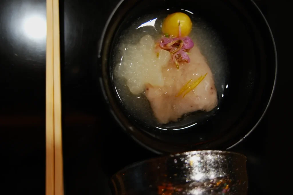 A delicate Japanese dish in a black bowl features fish, daikon radish, a yellow cherry tomato, and pink flower petals in clear broth, creating an elegant and serene presentation. Chopsticks rest beside the bowl.