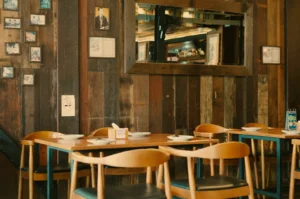 A cozy restaurant interior featuring wooden walls, several framed pictures, and a large mirror. There are empty wooden tables and chairs set for dining.
