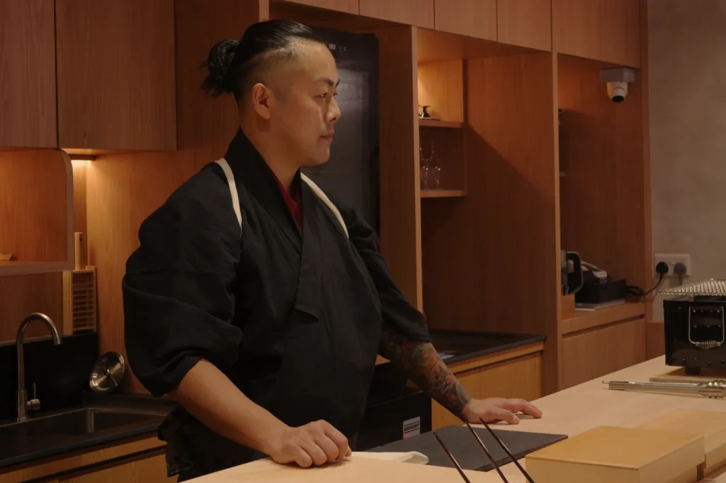 Chef Masa in a traditional Japanese chef's uniform stands at a kitchen counter, looking thoughtfully to the side.