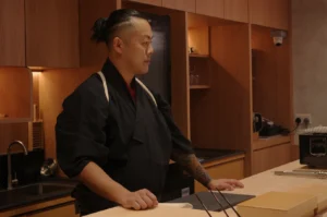 Chef Masa in a traditional Japanese chef's uniform stands at a kitchen counter, looking thoughtfully to the side.