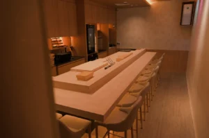 A minimalist kitchen with a long wooden dining table and stools, featuring a sushi preparation area in the middle.