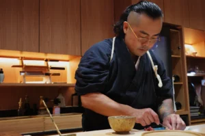 Chef Masa preparing food in the kitchen, wearing a traditional black chef's outfit and glasses.