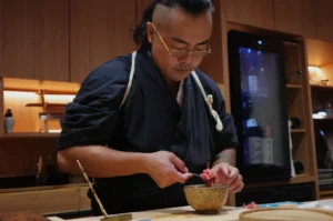 Chef Masa preparing food in a kitchen setting, wearing traditional attire and glasses, focused on his task.