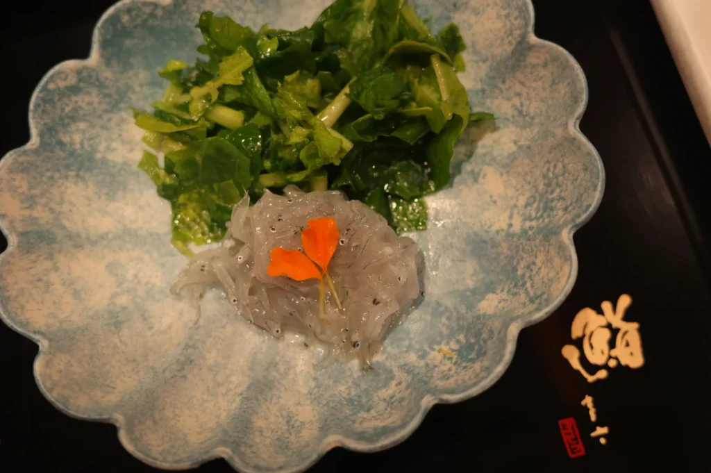 A blue scalloped dish containing transparent jelly-like substance garnished with an orange flower, alongside a serving of green leafy vegetables.