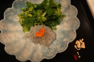 A blue scalloped dish containing transparent jelly-like substance garnished with an orange flower, alongside a serving of green leafy vegetables.