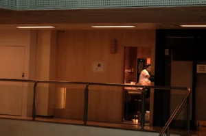 A dimly lit hallway with a railing, showing a doorway to a kitchen area where Chef Masa ina white shirt and an apron is preparing food.