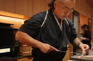 Chef Masa in a black kimono prepares sushi, skillfully slicing fish on a cutting board, with a kitchen setting in the background.