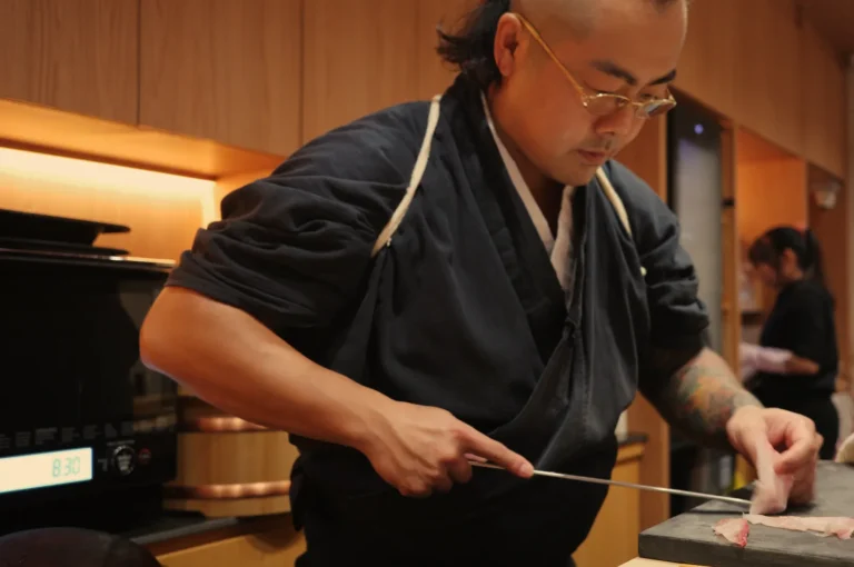 Chef Masa in a black kimono prepares sushi, skillfully slicing fish on a cutting board, with a kitchen setting in the background.