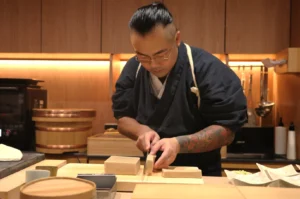 Chef Masa in a traditional attire is meticulously slicing ingredients on a wooden countertop in a warm, well-lit kitchen.