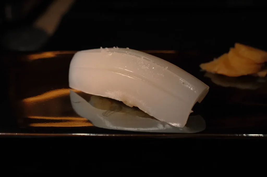 A piece of sushi featuring a slice of white squid, served on a dark plate with a small portion of pickled ginger beside it.