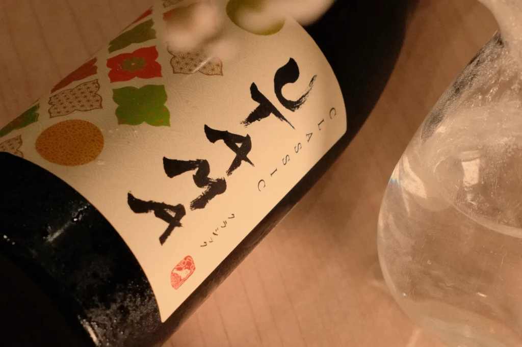 A close-up of a bottle of wine featuring a decorative label with stylized text in black and colorful shapes. A glass partially filled with liquid is next to the bottle.