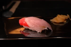 A piece of sushi on a dark plate, with a small mound of pickled ginger beside it.