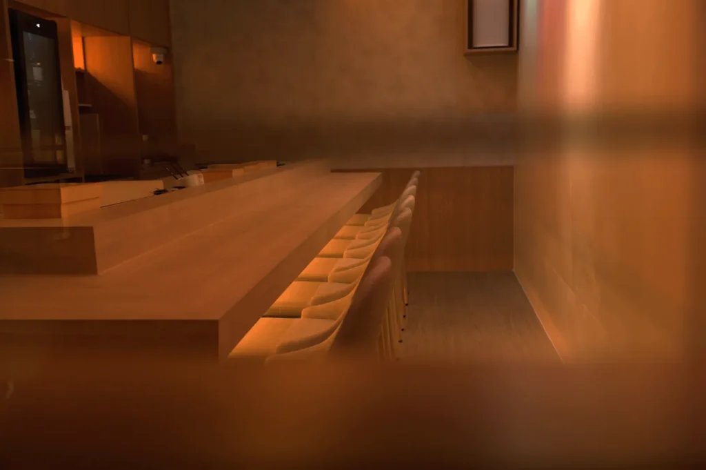 A minimalist restaurant interior featuring a wooden bar and seating, with warm lighting and a soft, muted atmosphere.