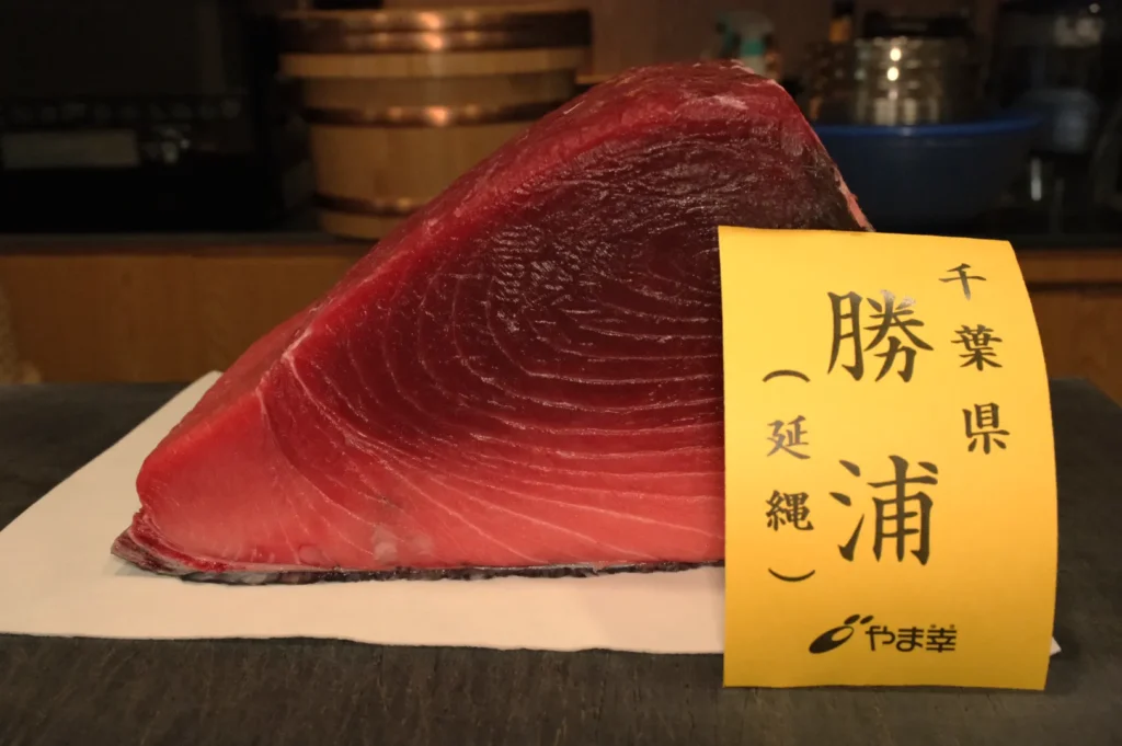 A large, triangular cut of tuna with a yellow label reading "千葉県 勝浦 一延縄."
