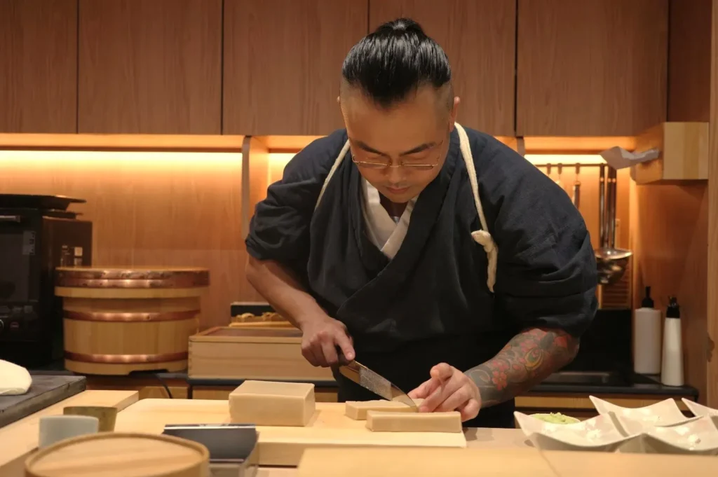 Chef Masa in traditional attire preparing food in a kitchen, focused on cutting an ingredient with a knife.