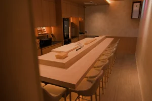 A modern, minimalist sushi restaurant interior with a long wooden counter, seating for diners, and simple decor.