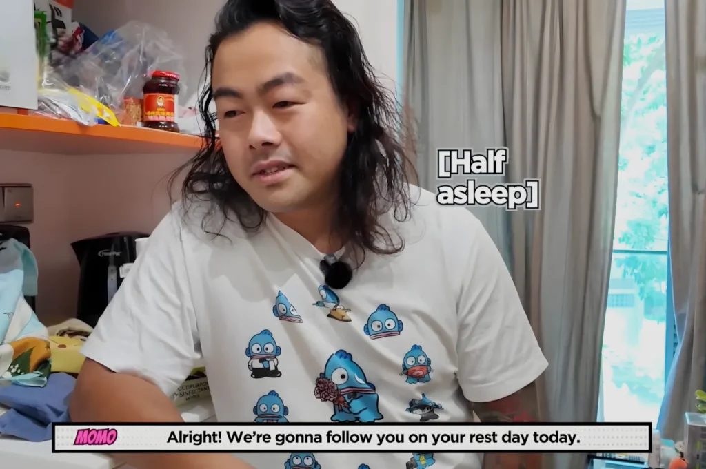 Chef Masa with long hair wearing a shirt featuring cartoon characters, sitting at a table with various items around him, looking slightly drowsy. Text overlay says "[Half asleep]" and another text reads, "Alright! We’re gonna follow you on your rest day today.”