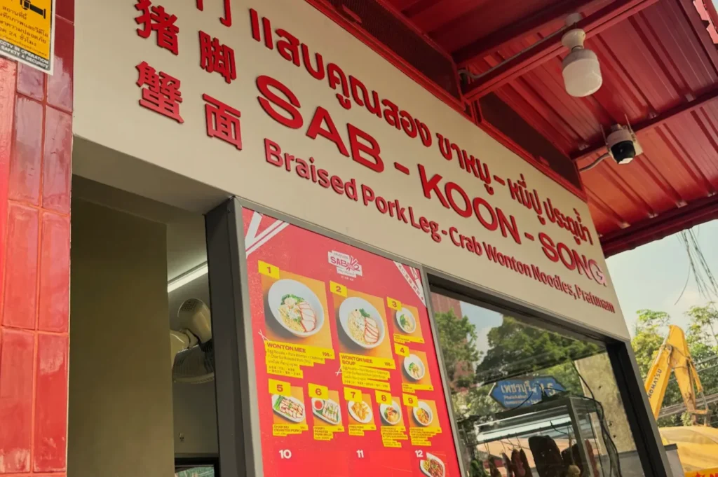 A restaurant sign for "SAB-KOON-SONG" featuring the text "Braised Pork Leg Crab Wonton Noodles Restaurant" along with a menu display.
