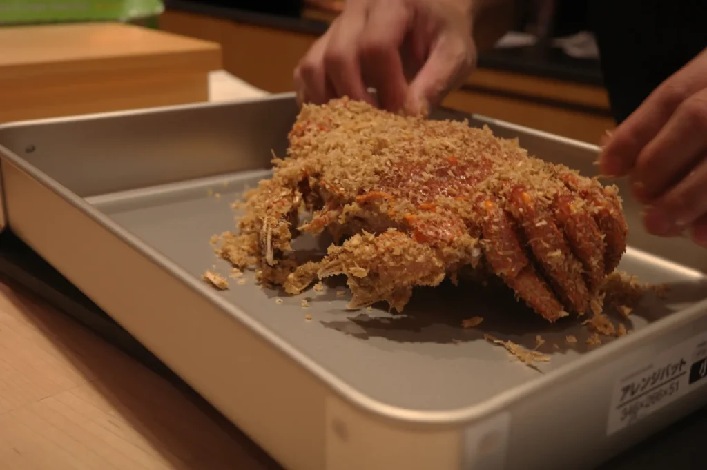A person preparing a dish with a breaded crab on a baking tray.