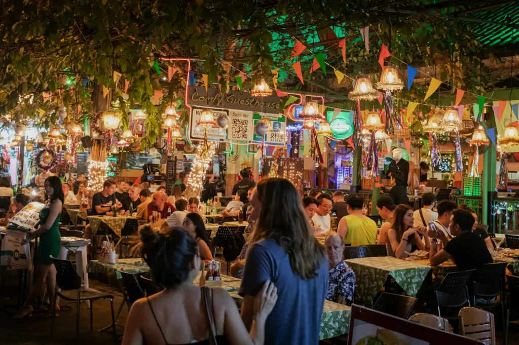 A vibrant and lively outdoor dining scene at night, with colorful lights, decorations, and people enjoying their meals at tables under a canopy of greenery.
