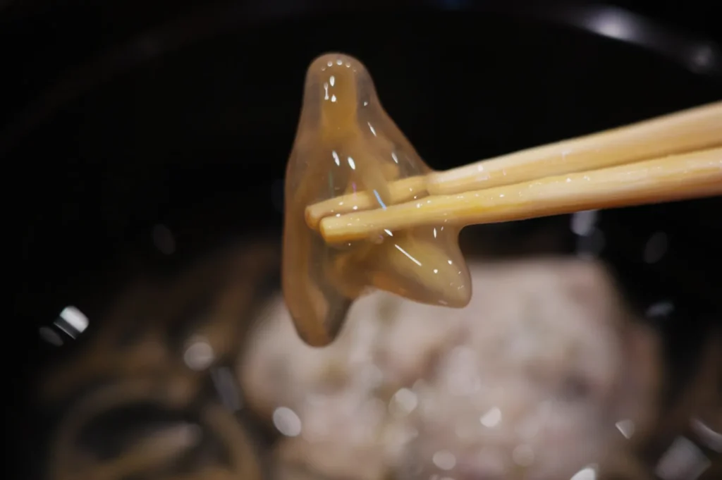 A pair of chopsticks holding a translucent, gelatinous piece of food above a dark bowl of broth.