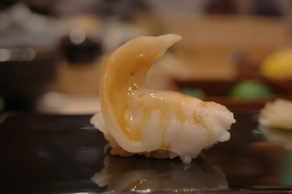 A piece of sushi on a black plate, featuring a slice of sushi topped with a pale colored seafood.