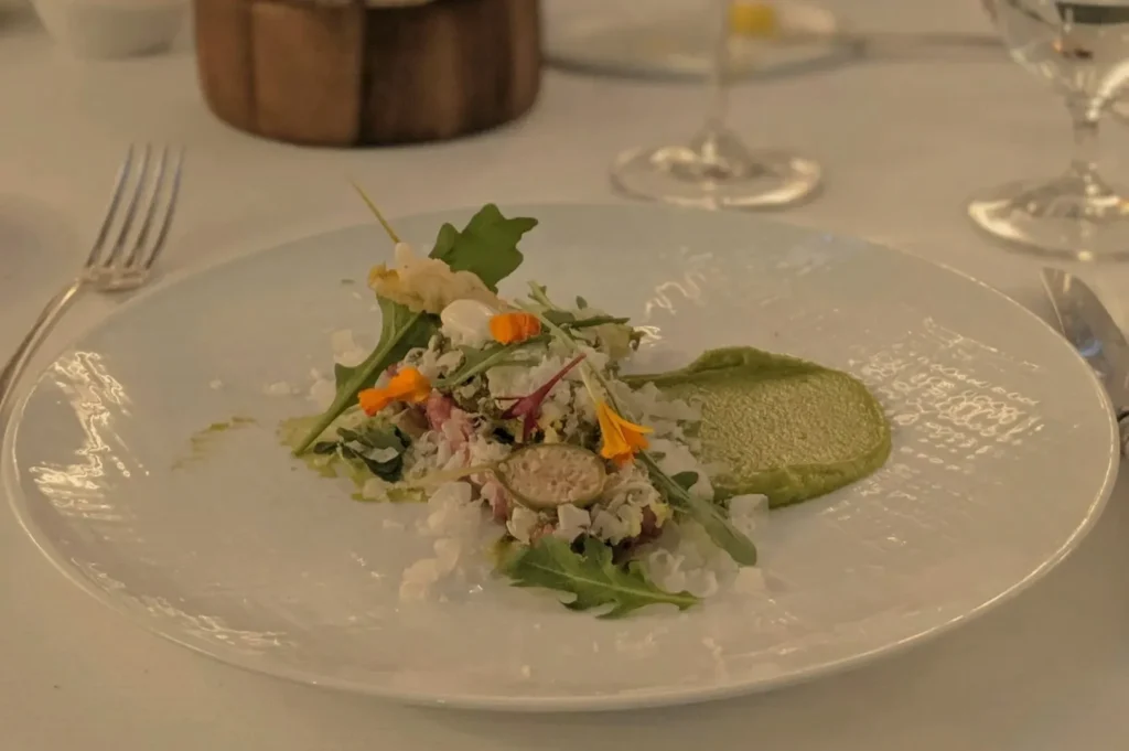 A beautifully plated dish featuring a mix of greens, delicate garnishes, and a smooth green sauce, presented on a white plate.